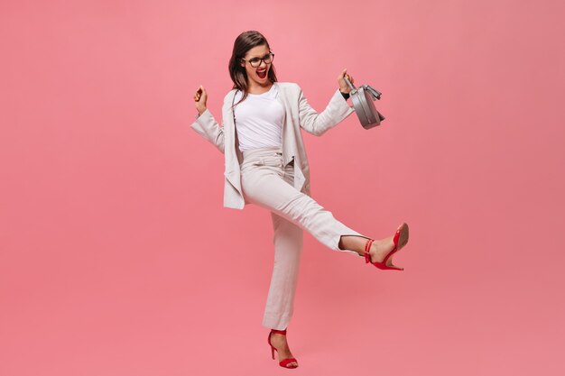 Senhora ativa de terno posando com bolsa em fundo rosa. Mulher jovem emocional se divertindo na câmera de salto alto vermelho.