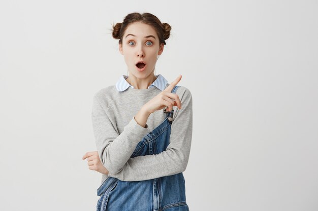 Senhora atônita em macacão jeans elegante, apontando o dedo sobre a parede branca. Professora na moda mostrando algo chocante com a boca aberta. Copie o espaço