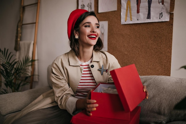 Foto grátis senhora atraente com roupa de estilo parisiense abre caixa vermelha jovem feliz em boina vermelha brilhante e casaco bege estiloso posando
