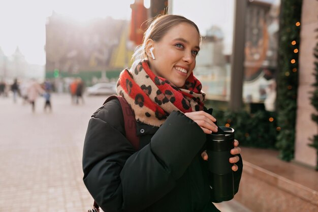 Senhora bonita elegante com sorriso adorável está bebendo café e ouvindo música e gostando de caminhar na rua ensolarada