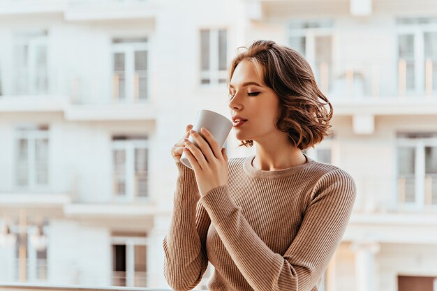 Senhora branca inspirada com penteado encaracolado, bebendo chá. Mulher jovem e linda desfrutando de café na manhã fria de outono.