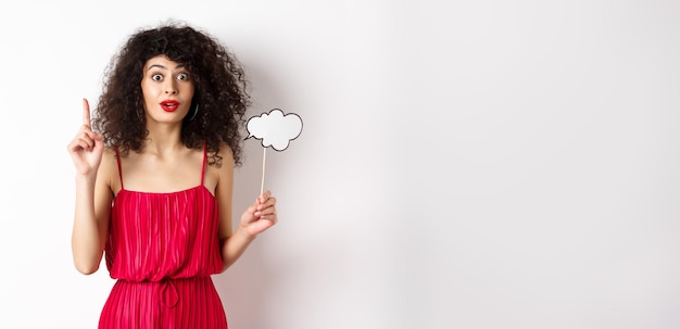 Foto grátis senhora caucasiana bonita em vestido vermelho segurando nuvem de comentários e levantando o dedo lançando uma ideia e sorrindo