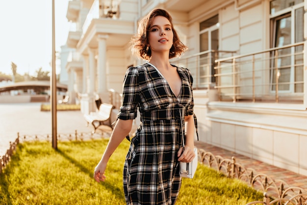 Senhora caucasiana encantadora em vestido elegante, posando na cidade. ainda bem que mulher branca com cabelos ondulados, andando na rua em um dia ensolarado.