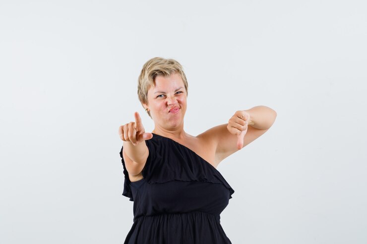 Foto grátis senhora charmosa de blusa preta apontando para a câmera enquanto mostra o polegar para baixo e parece descontente