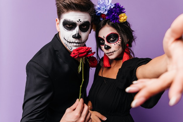 Senhora com coroa de rosas e brincos vermelhos tira selfie enquanto jovem com arte de rosto para o Halloween segura rosa.