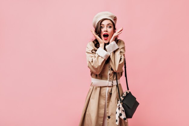 Senhora com lábios vermelhos está olhando para a câmera surpresa. Mulher com casaco elegante posando com bolsa crossbody no fundo rosa.