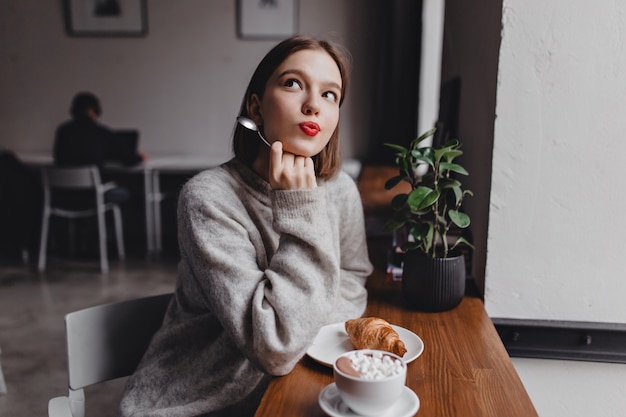 Senhora com roupa enorme cinza posando sonhadoramente no café. Retrato de jovem à mesa com croissant e cappuccino.