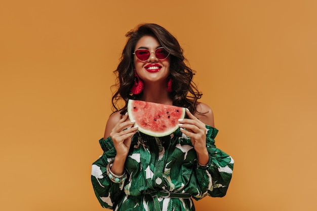 Senhora da moda sorridente com penteado moderno encaracolado em brincos vermelhos e roupas elegantes verdes posando com melancia em pano de fundo laranja