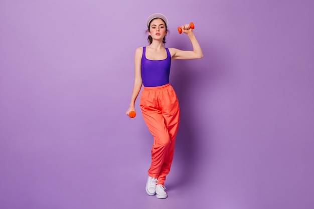 Senhora de blusa esportiva roxa e calça laranja fazendo exercícios para as mãos com halteres vermelhos