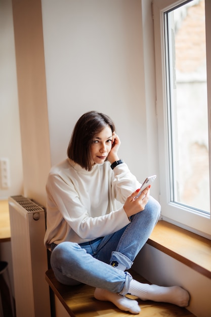 Senhora de cabelos curtos em jeans azul, sentada no peitoril da janela e escrever mensagem no smartphone em casa