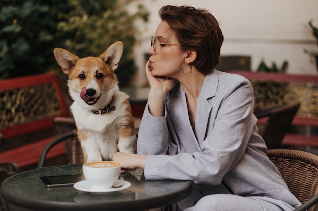 Senhora de cabelos curtos gosta de café no café e olha para o cachorro. Mulher encantadora com jaqueta cinza descansando com corgi do lado de fora