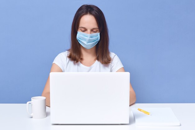 senhora de cabelos escura, vestindo camiseta casual e máscara médica que trabalha na frente da tela do colo superior, parece concentrado, fêmea estudante fazendo tarefa universitária on-line, educação.