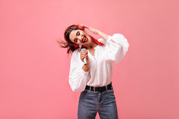 Senhora de cabelos escuros em jeans e blusa branca elegante ouve música em fones de ouvido e canta no microfone no fundo isolado.