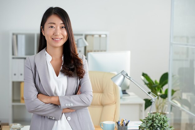 Senhora de negócios coreano alegre posando no escritório com os braços cruzados