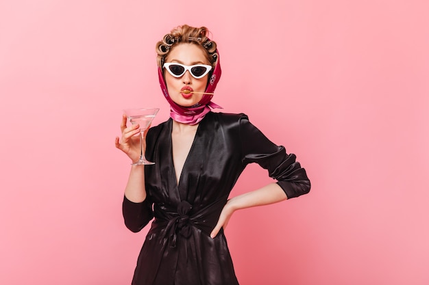 Senhora de óculos escuros posando na parede rosa, segurando uma taça de martini e comendo azeitona