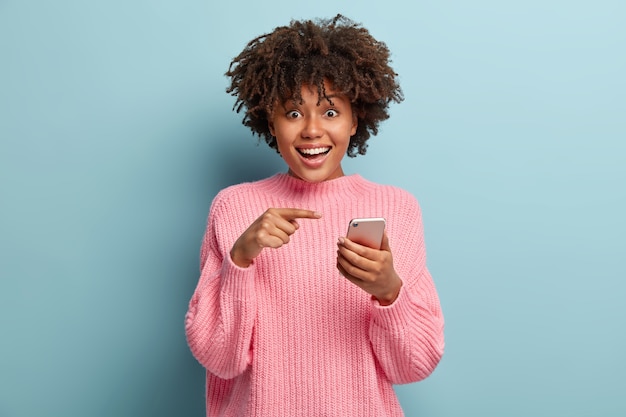 Senhora de pele escura, positiva e surpresa aponta para dispositivo de telefone inteligente, sugere ter uma conversa no bate-papo em grupo, feliz por receber muitas mensagens de parabéns na caixa de correio, tem olhar radiante