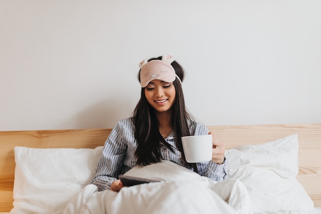 Senhora de pijama e máscara de dormir segura uma xícara de chá e lê enquanto está deitada na cama