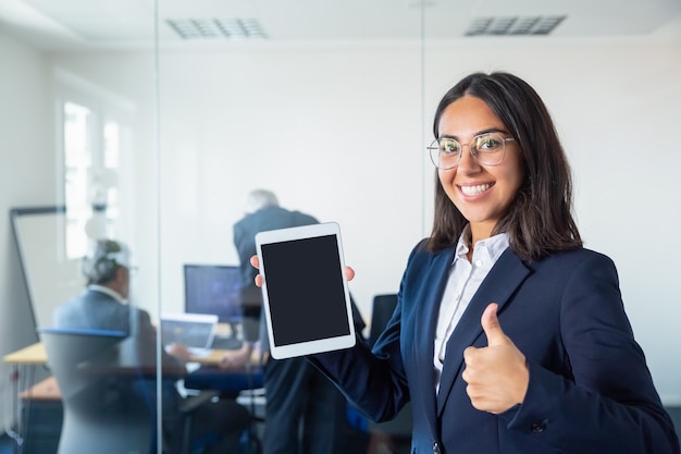 Senhora do escritório feliz mostrando a tela do tablet em branco, fazendo um gesto, olhando para a câmera e sorrindo. copie o espaço. conceito de comunicação e publicidade