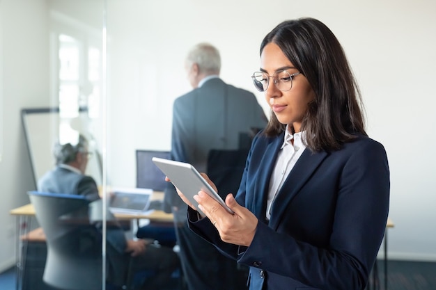 Senhora do escritório focada em copos usando tablet enquanto dois empresários maduros discutindo o trabalho atrás da parede de vidro. copie o espaço. conceito de comunicação