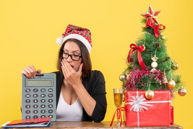 Senhora encantadora chocada de terno com chapéu de Papai Noel e óculos mostrando calculadora no escritório em amarelo isolado