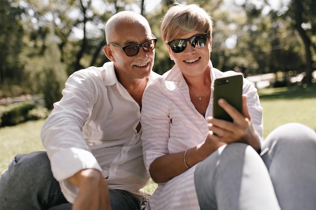 Senhora encantadora com penteado curto em óculos de sol e blusa rosa olhando para smartphone e sorrindo com homem de cabelos grisalhos na camisa branca no parque