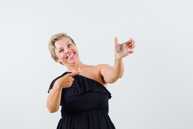 Senhora encantadora de blusa preta posando como se estivesse segurando um telefone enquanto aponta para ele e parece positiva