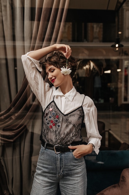 Senhora encantadora de camisa branca com renda preta e calça jeans, sorrindo no café. Mulher com lábios brilhantes e cabelo curto segura uma flor dentro.