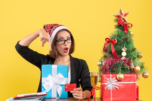 Senhora encantadora de terno com chapéu de papai noel e óculos apontando para presente e cartão de banco no escritório