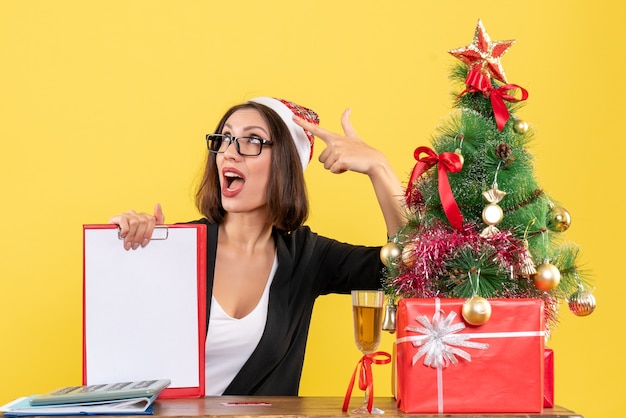 Senhora encantadora de terno com chapéu de papai noel e óculos mostrando documento, sentindo-se chocada no escritório