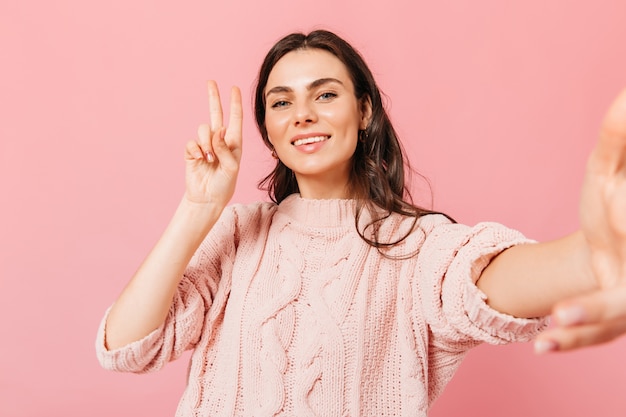 Senhora encantadora de vestido de malha faz selfie no fundo rosa. Mulher de cabelos escuros em alto astral, mostrando sinal de paz.