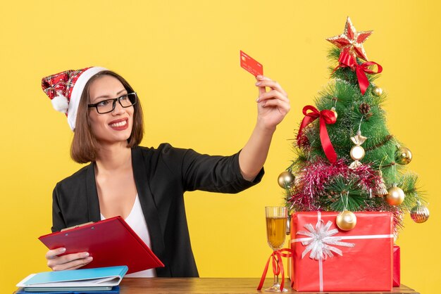 Senhora encantadora orgulhosa de terno com chapéu de Papai Noel e óculos mostrando o cartão do banco no escritório em amarelo isolado