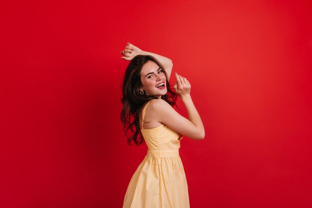 Senhora encaracolada travessa está dançando na parede vermelha. Morena de vestido amarelo sorri sinceramente e gosta de sessão de fotos.