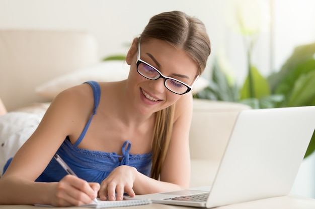 Senhora escrevendo no caderno ao usar o laptop em casa