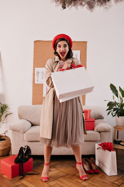 Senhora espantada com boina vermelha e pose bege com sacola de compras branca. Mulher surpreendida com chapéu brilhante e casaco de outono Olha para a câmera no apartamento.