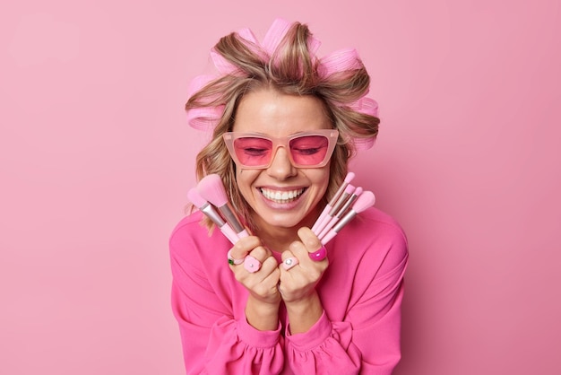 Foto grátis senhora feliz aplica rolos de cabelo para fazer penteado usa óculos escuros da moda blusa de areia segura pincéis cosméticos que vão aplicar maquiagem sorrisos amplamente isolados sobre parede rosa tratamentos de beleza femininos