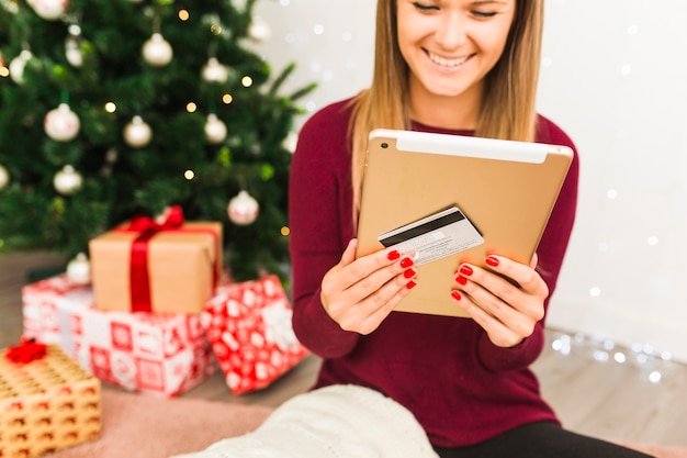 Senhora feliz com tablet e cartão de plástico perto de caixas de presente e árvore de Natal