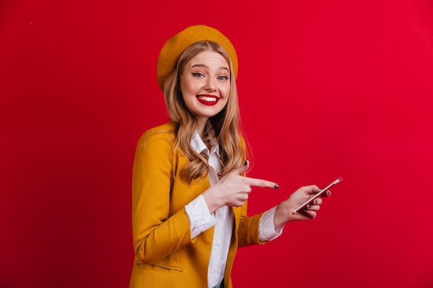 Senhora francesa alegre apontando com o dedo para o smartphone. Menina loira sorridente de boina em pé na parede vermelha