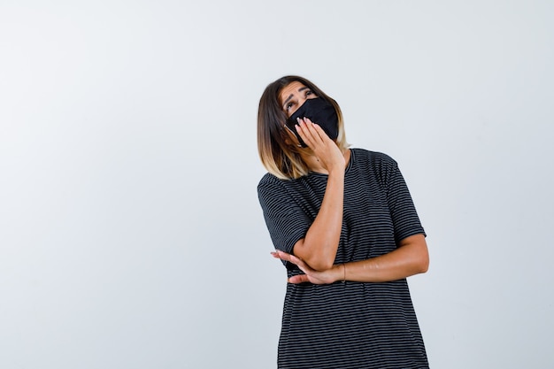 Senhora inclinando a bochecha na palma da mão em um vestido preto, máscara médica e olhando pensativa, vista frontal