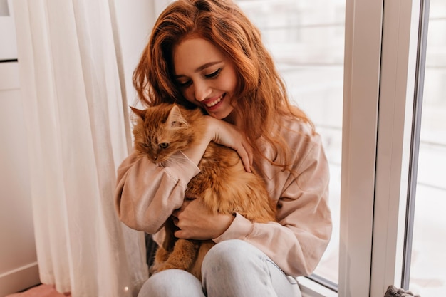 Senhora jocunda com cabelo ondulado posando com seu animal de estimação Retrato interior de menina europeia brincando com gato em casa