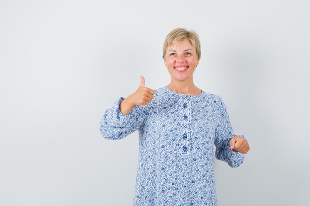 Senhora loira aparecendo o polegar na blusa estampada e parecendo alegre, vista frontal.