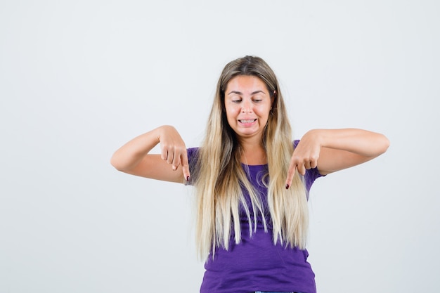 Senhora loira apontando para baixo em t-shirt violeta e olhando curiosa, vista frontal.