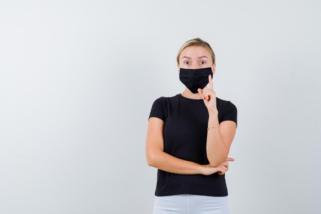 Senhora loira apontando para cima em camiseta preta, máscara preta e parecendo cuidadosa isolada