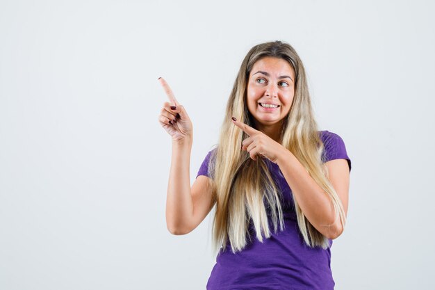 Senhora loira apontando para o canto superior esquerdo em uma camiseta violeta e olhando alegre. vista frontal.