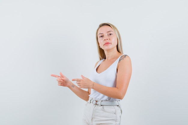 Foto grátis senhora loira apontando para o lado em camiseta, calças e parecendo confiante, vista frontal.