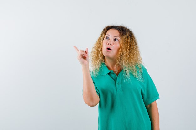 Senhora loira com cabelo encaracolado apontando para o canto superior esquerdo em uma camiseta verde e parecendo curiosa, vista frontal.