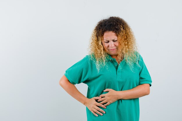 Senhora loira com cabelos cacheados, sofrendo de dor abdominal em uma camiseta verde e parecendo indisposta. vista frontal.