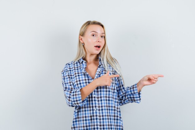 Senhora loira com camisa xadrez apontando para o lado direito e parecendo feliz