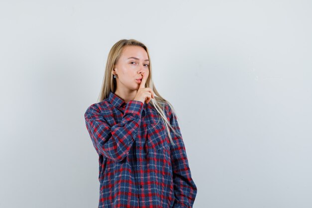 Senhora loira de camisa casual, mostrando o gesto de silêncio e olhando confiante, vista frontal.