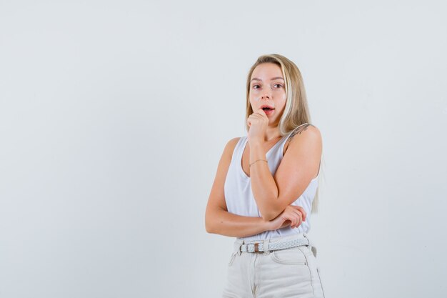 Senhora loira de camiseta, calça em pé em pose pensativa e parecendo espantada