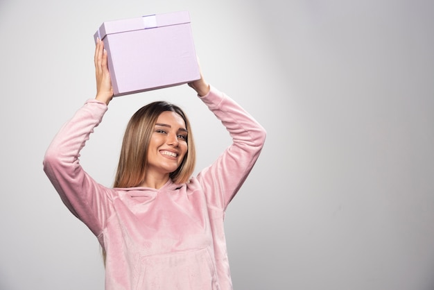 Senhora loira em moletom rosa segura uma caixa de presente na cabeça e balançando.